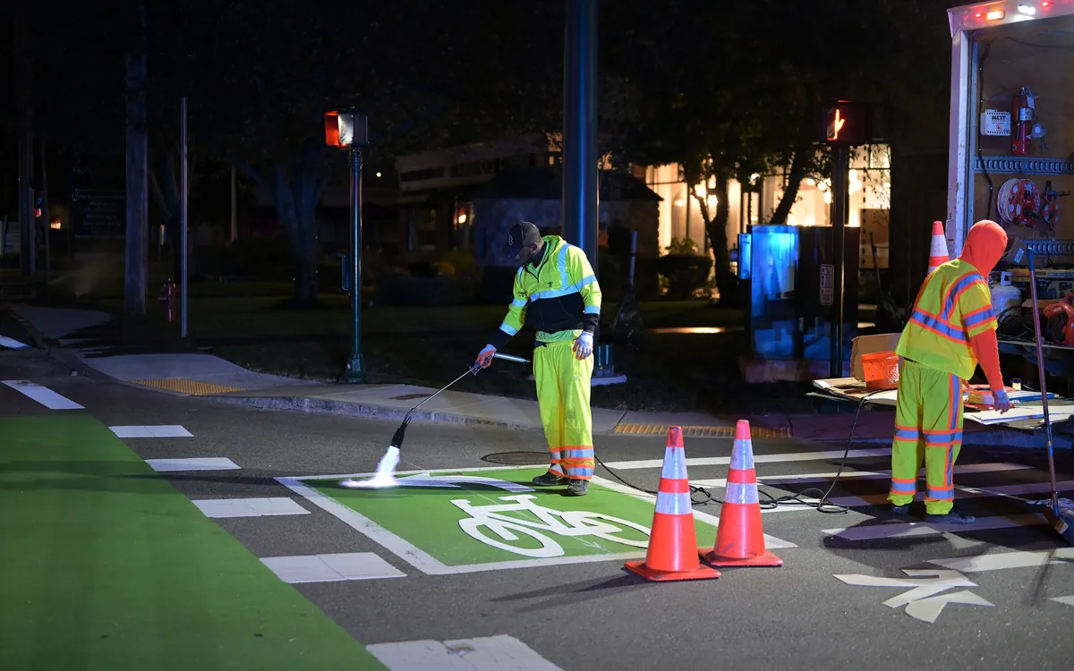 Two workers laying road markings at night with a blowtorch, traffic cones around them.